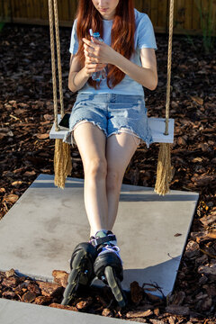 Red haired tenage girl wearing roller skates sits outdoors and drinks water after skating in warm evening light. Concept of hydration after exercise, healthy habit and active teenage lifestyle.