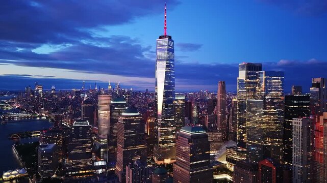 Aerial panorama at night, New York City. Aerial perspective of the illuminated skyscraper spire, surrounding financial district buildings and waterfront at dark.