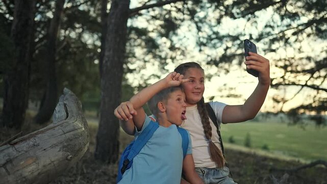 Children sit together using tablet in forest. Boy and girl laugh while taking selfie. Sibling shares tablet, sibling shares smile. Forest gives space. Selfie repeats, children enjoy selfie again.