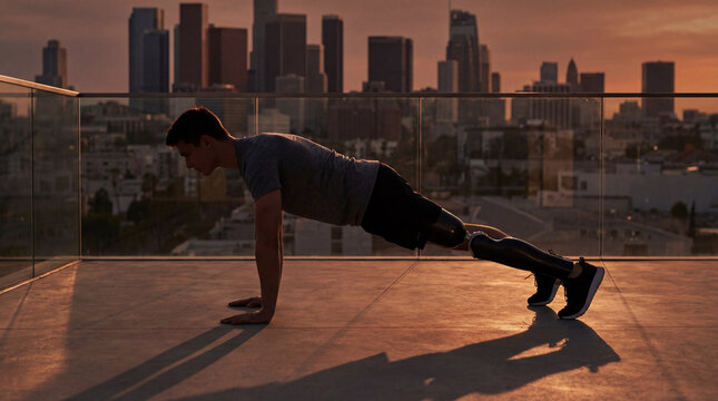 Man with prosthetic legs doing push-ups on rooftop at sunset with city skyline.