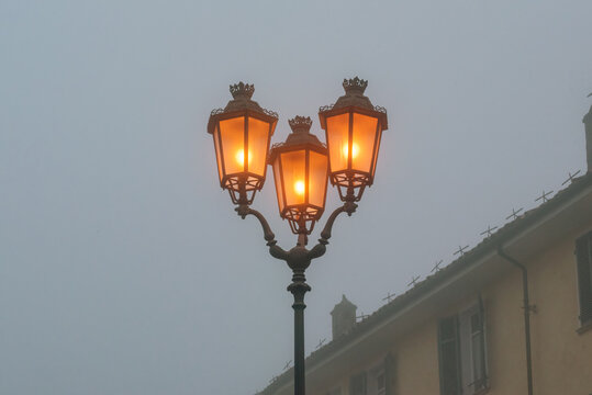 A vintage-style triple-headed street lamp glows warmly against a dense, melancholic fog, casting an inviting amber light that cuts through the grey, atmospheric silence of an early morning urban scene