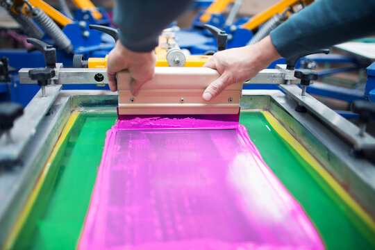 Silk screen printing technology. Serigraphy and Color paints applying to fabric. Plastisol paint and squeegee in worker hands