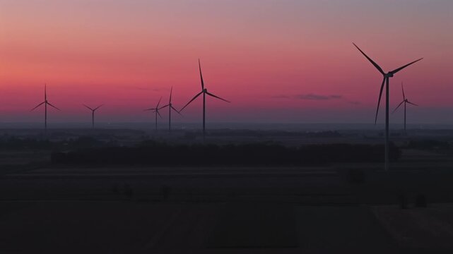 A wind farm at dawn, with towering wind turbines rotating gracefully in the breeze. Red aviation warning lights flash atop the towers, while the turbines harness the wind to produce energy.