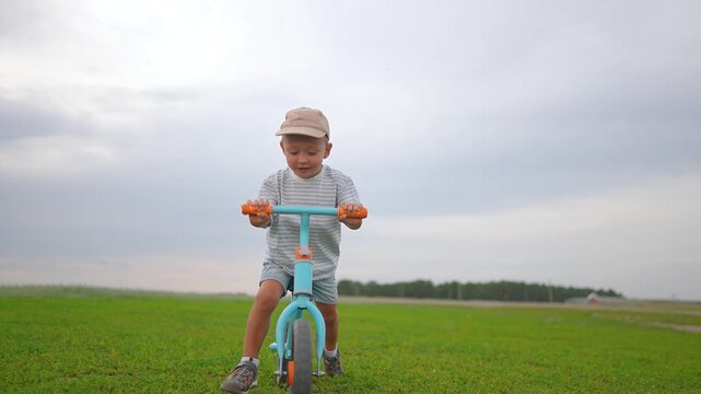 Smiling kid rides bike in sunny park. Happy child riding bicycle through green grass. Boy enjoys outdoor play. Kid riding bike on path. Toddler smiles while riding bicycle in outdoor park again.