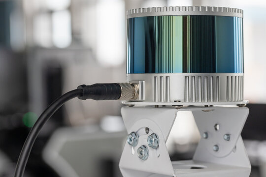 LiDAR sensor mounted on a metal bracket with cable connection, photographed in a workshop setting. Close-up shows precision housing, cooling fins and connector used for 3D mapping and robotics