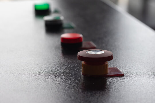 Close-up of a red emergency stop button mounted on a textured industrial control panel, with a blurred row of green and red indicator buttons receding into the background