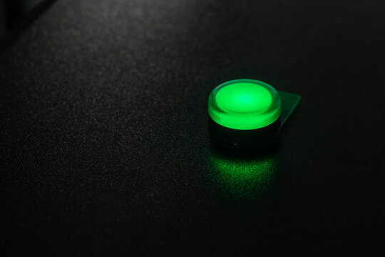 Small green illuminated button glowing on a textured black surface, casting a soft reflection; a minimal electronic power indicator conveying activation, status, technology and modern interface design