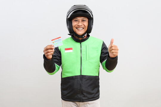 Cheerful Asian online motorbike taxi driver holding ID card and driving license while giving thumbs up, isolated on grey background.