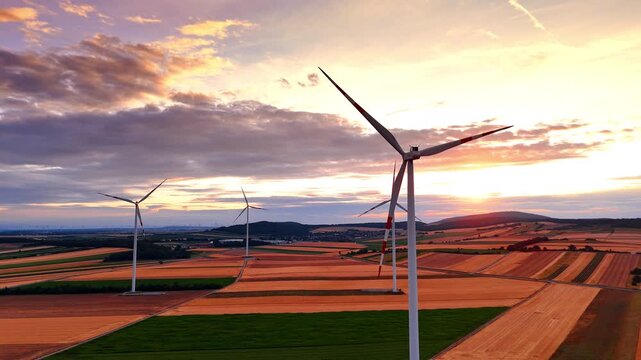 Wind turbines spanning farmland beneath glowing sunset clouds. Elevated view shows four white towers, patchwork fields, hills and backlit sky in warm evening light.