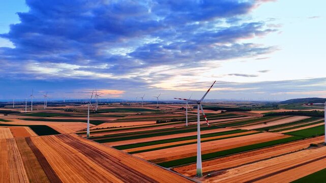 Wind turbines distributed across striped farmland beneath blue clouds. Aerial panorama centers two white towers with red blade markings and rows of distant generators.