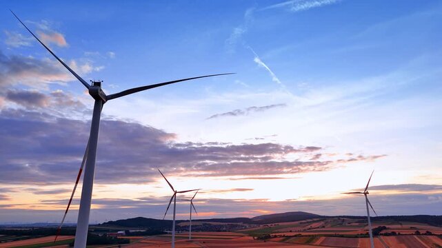 Close wind turbine above fields under blue evening sky. Low aerial angle shows a cropped white tower and three distant turbines, with clouds and contrails.