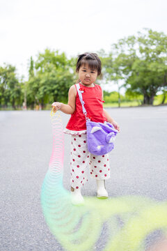 Adorable Asian Girl Playing with a Rainbow Slinky Spring Toy Outdoors,A cute young Asian girl standing in a park, holding and playing with a vibrant, colorful rainbow-colored slinky spring toy.
