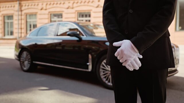 Professional chauffeur wearing black suit and gloves standing with clasped hands near luxury black sedan. Footage is ideal for luxury transport and VIP concierge service commercials
