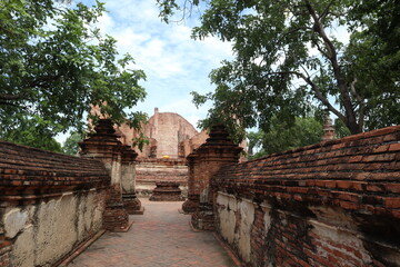 Fototapeta premium Ancient Temple Path: A stone pathway winds through ancient brick walls toward a weathered temple ruin, framed by lush green trees under a cloudy sky.