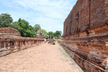 Fototapeta premium Ancient Brick Temple Wall Pathway: A perspective shot of a weathered brick pathway leading alongside a massive ancient temple wall, with remnants of structures and lush greenery in the background.