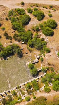 Vertical video. Balat, Turkey. Aerial drone view over the ancient Roman ruins of Miletus, revealing the Nymphaeum fountain, Capito Baths, and North Market amid sun-baked earth and scattered olive gro