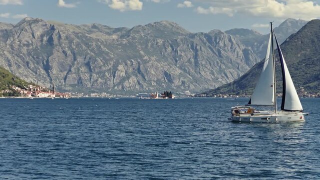 aerial view toward the island of our lady of the rocks and the town of perast in the bay of kotor, framed by steep mountains and fair weather clouds, montenegro