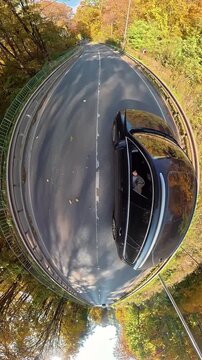 Creative 360 degree tiny planet view of black car driving on asphalt road, golden autumn forest surrounding curved highway in sunny day