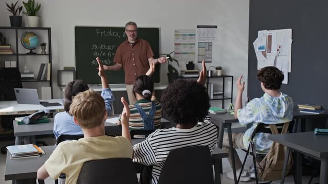 Middle aged Caucasian male teacher explaining math equations on blackboard to multiethnic teenagers raising hands in classroom. Footage for high school promotions and e-learning campaigns