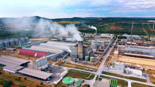 Thick white smoke comes out of the pipes in the industrial area. Green nature landscape in the backdrop.