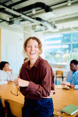Fototapeta na wymiar Woman smiling and holding glasses, standing in modern office with diverse colleagues collaborating at a long table