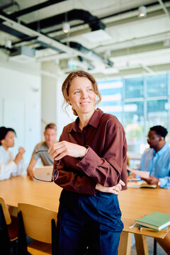 Professional woman standing with glasses in hand, smiling, leading a team meeting in a modern open-plan office space