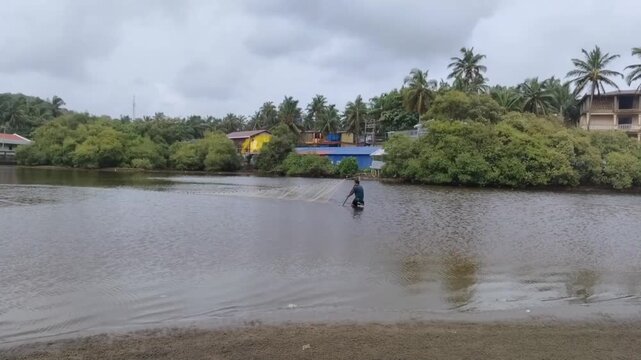Daytime outdoor shot of an Asian male fisherman casting fish net in Konkan and Goa in India on cloudy day. Locals fishing in a lake trying to catch a fish with tropical trees  and houses in background