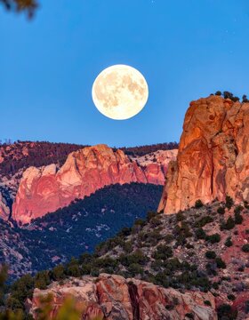 A full moon hangs in the twilight sky over rugged, sun-kissed mountains.