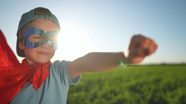 Boy wears superhero mask with red cape standing in summer field. Happy child raises fist pretending hero. Mask covers boy face. Cape flutters outdoor. Boy plays superhero enjoying fun adventure field.