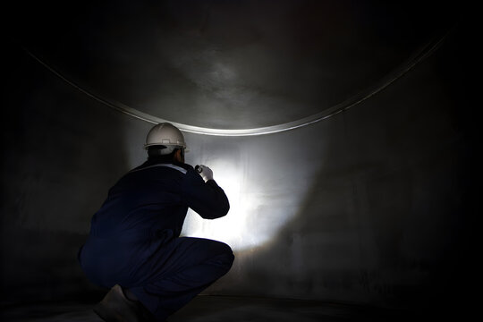 Industrial inspector in white hard hat and blue coverall examining rusted metal tank interior wall with flashlight beam, confined space internal tank inspection and corrosion assessment concept