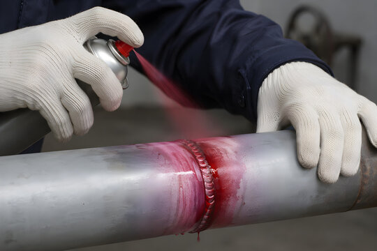 Industrial technician applying red liquid penetrant dye spray on metal weld seam for non-destructive testing NDT inspection quality control in chemical manufacturing plant facility