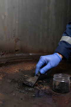 Gloved industrial worker hand using metal spatula collecting dark sediment sample from corroded storage tank floor into glass jar, confined space contamination analysis procedure