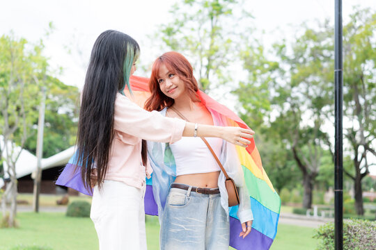 Loving young Asian lesbian couple sharing rainbow pride flag in green park. Beautiful LGBT women looking at each other with tenderness, celebrating gender diversity and equality concept.