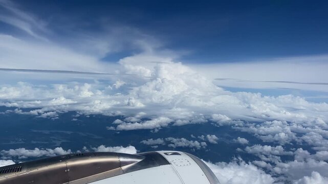 Wide panoramic view from airplane window showing curved wing, deep blue sky, fluffy cumulus clouds, and ocean below during daytime.