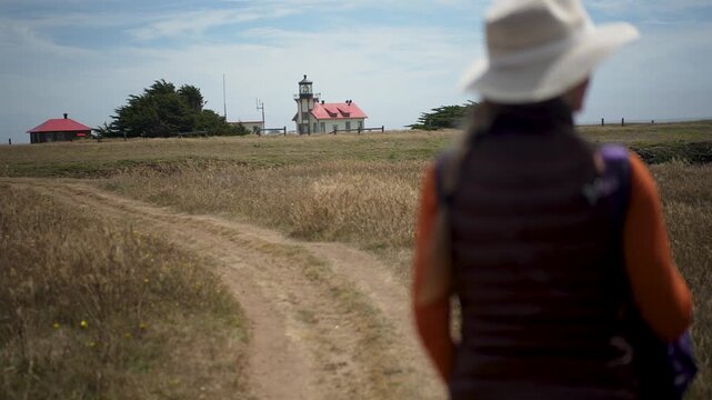 Mature woman walks along trails heading to Point Cabrillo Lighthouse in Mendocino California near the Pacific Ocean on a sunny day.