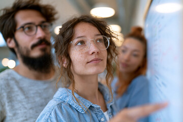 Obraz premium Young team gathers around whiteboard in bright office. Focused members collaborate, brainstorming ideas. Woman gestures while others listen intently. Warm lighting enhances professional