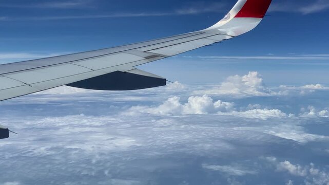View from airplane wing flying high above a thick layer of white and gray clouds under a bright blue sky, with red and white wingtip marking visible.