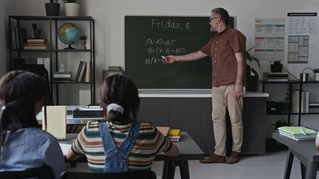 Rack focus shot of middle-aged male teacher explaining math equations on blackboard to schoolgirls in classroom. Footage for educational marketing, school promo videos, and e-learning platforms