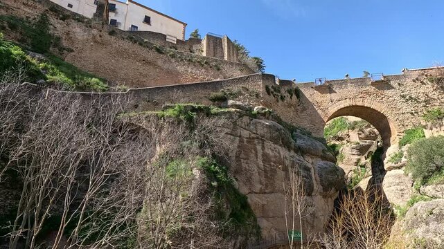 Wide low-angle shot of a deep rocky gorge in Ronda, Spain, with a massive stone arch bridge spanning the chasm and multi-story buildings perched on tiered retaining walls along the cliff face.
