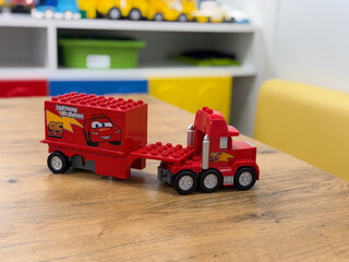 Fototapeta premium Red toy truck with a cartoon race car trailer on a wooden table in a playroom, with shelves and colorful bins in the background