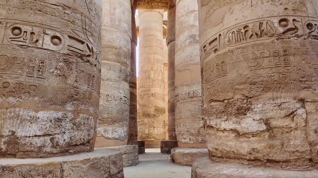 POV walking through Monumental stone columns inside the great hypostyle hall at karnak temple. Carved ancient egyptian hieroglyphs covering the massive pillars