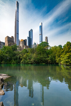 Scenic view from Central Park showcasing the tall and slender skyscrapers of Billionaires Row reflecting on the quiet surface of The Pond surrounded by trees.