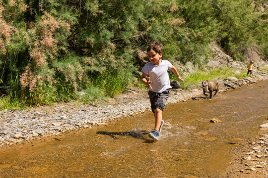 Young boy running through river with dog splashing water