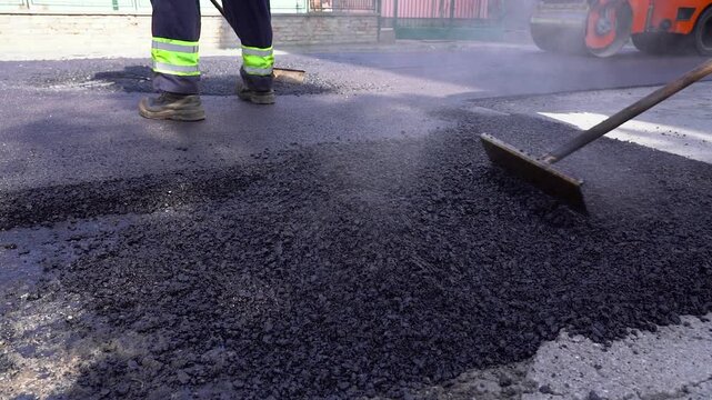 Worker Paving Hot Asphalt with Rake as Roller Compacts Road, Slow Motion.  Great for roadwork concepts, heavy machinery maintenance, and industrial development usage.