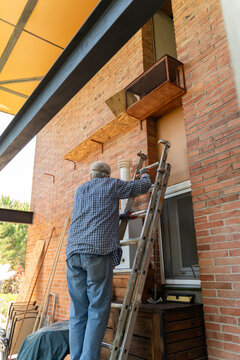 Older Man Works on Wooden Shelf on Brick Wall in Backyard 