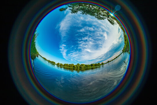Fisheye view of a river with blue water, clouds in the sky, and trees on the banks. HDR photo taken with a circular wide-angle lens