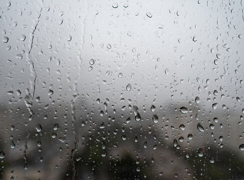 Rain drops on window glass during cloudy rainy weather with blurred city view in the background. Concept of autumn weather, sadness, melancholy, storm, climate, and urban rainy day background.