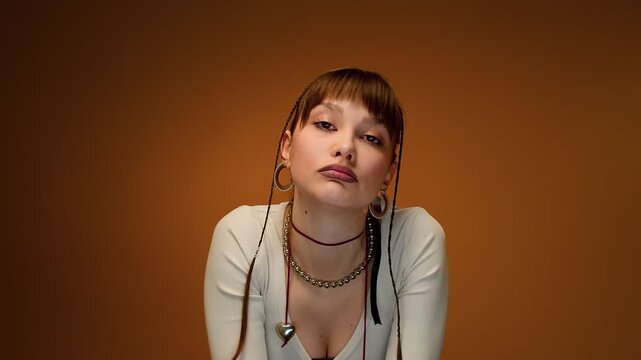 Intense Young Woman with Braided Hair and Blunt Bangs in Warm Studio Portrait Against Brown Backdrop