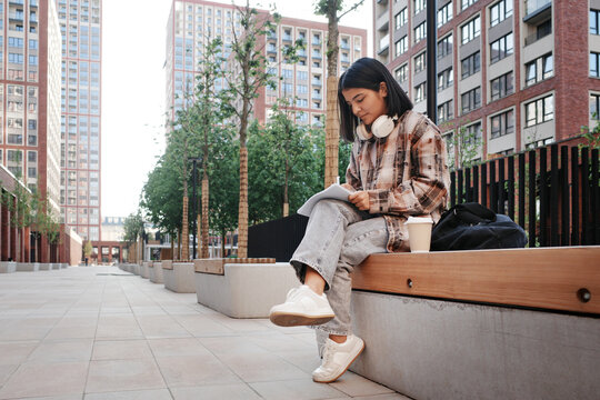 Student using phone on campus bench with coffee and headphones