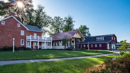 Obraz premium A red brick house with a porch sits next to a large green lawn. The barn and another building are nearby. The sky is clear, and sunlight shines on the scene.